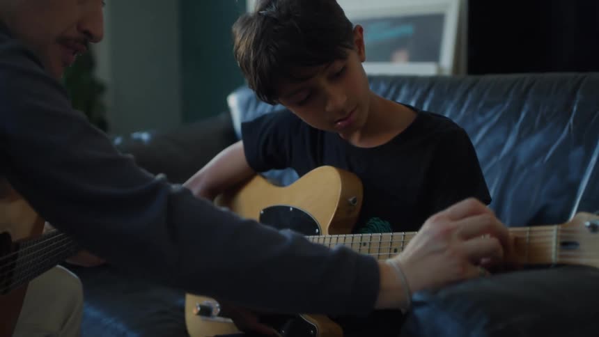 A man teaches a boy to play an acoustic guitar while sitting on a couch at home