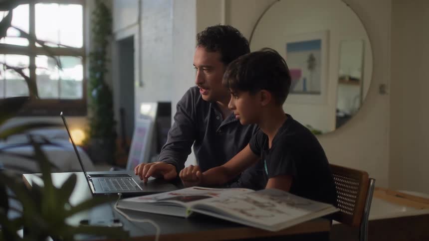 A father and son are looking at a laptop screen together with an open book on the table