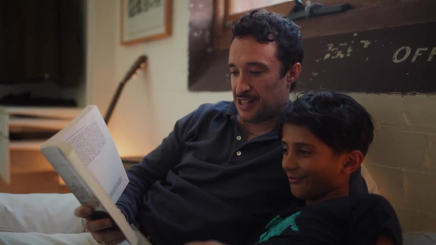 Father reads to his son from a book while they lie on the bed