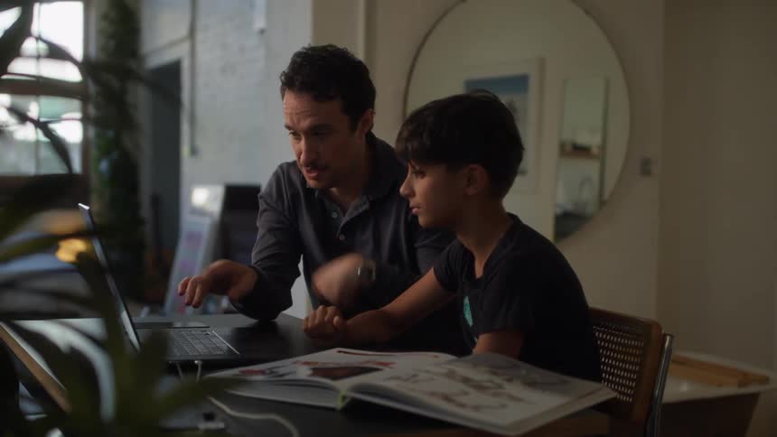 A father and son are seated at a table looking at a laptop screen together with an open book in front of them sharing a moment of learning and activity in a well lit modern home