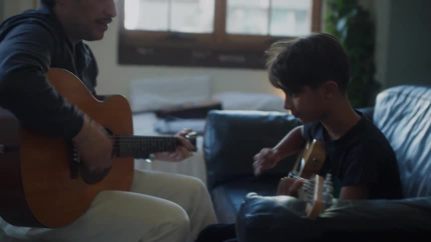 A man teaches a boy how to play guitar using an acoustic guitar while the child holds an electric guitar seated together on a couch at home