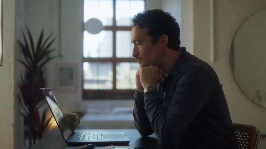 A man with dark curly hair sits at a desk looking intently at a laptop screen his chin resting on his clasped hands before he begins to type
