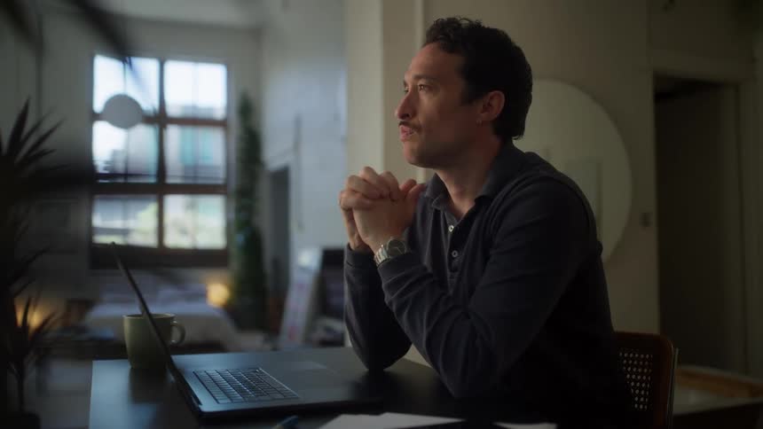 A man with dark curly hair sits at a desk looking at a laptop screen and typing with a mug and papers nearby in a softly lit room