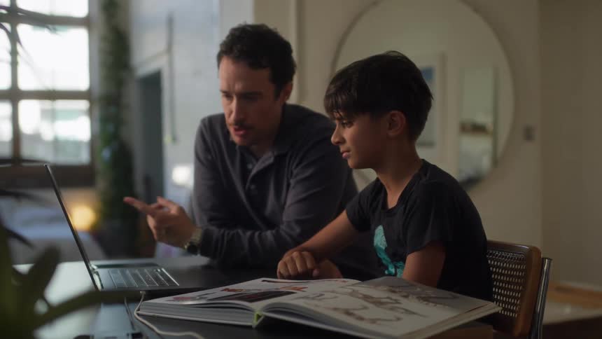 A father with a mustache and a dark shirt and son wearing a black t shirt are engaged in a learning activity at a table with the father pointing to something in an open book while the son looks on attentively