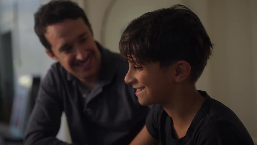 A father points to a laptop screen explaining something to his son who is listening attentively
