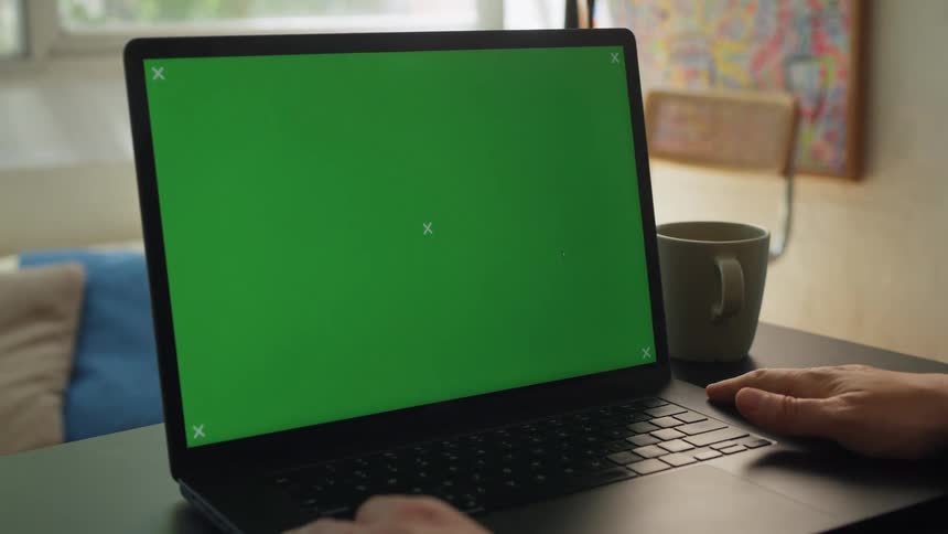 A laptop with a green screen display is positioned on a desk with a persons hands visible on the keyboard and a mug nearby