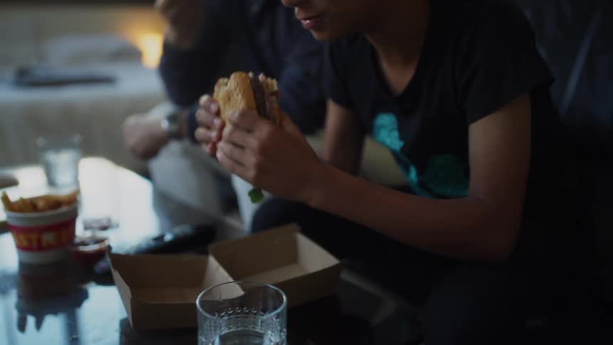 A father and son eat burgers with fries while watching television at home