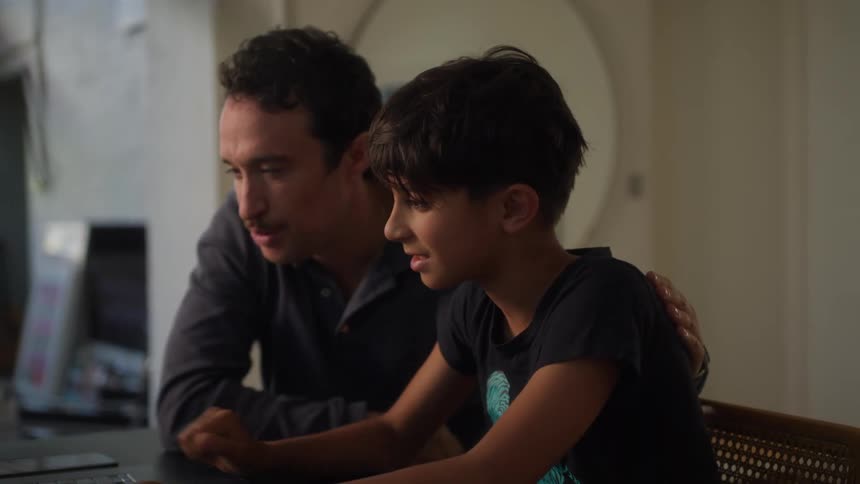 A man with a mustache and a dark shirt and a boy wearing a black t shirt look at a laptop and then give each other a high five