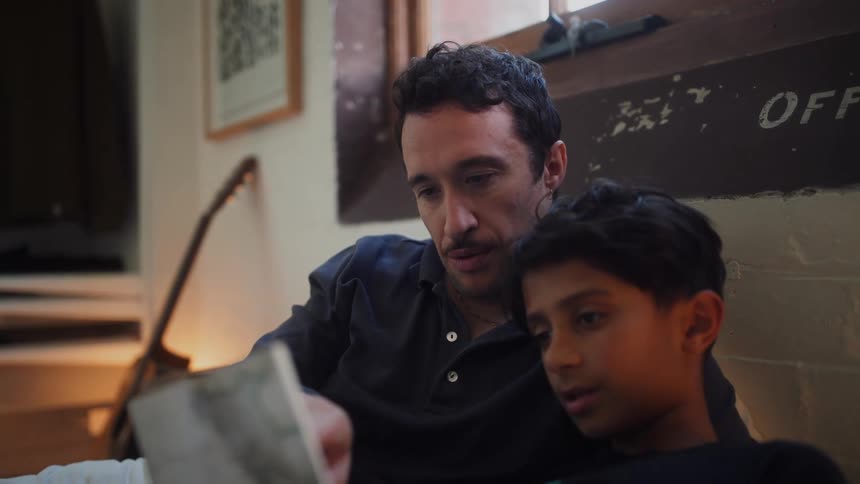 A man with a mustache a dark blue shirt white jeans and a boy wearing a black t shirt read a book together indoors