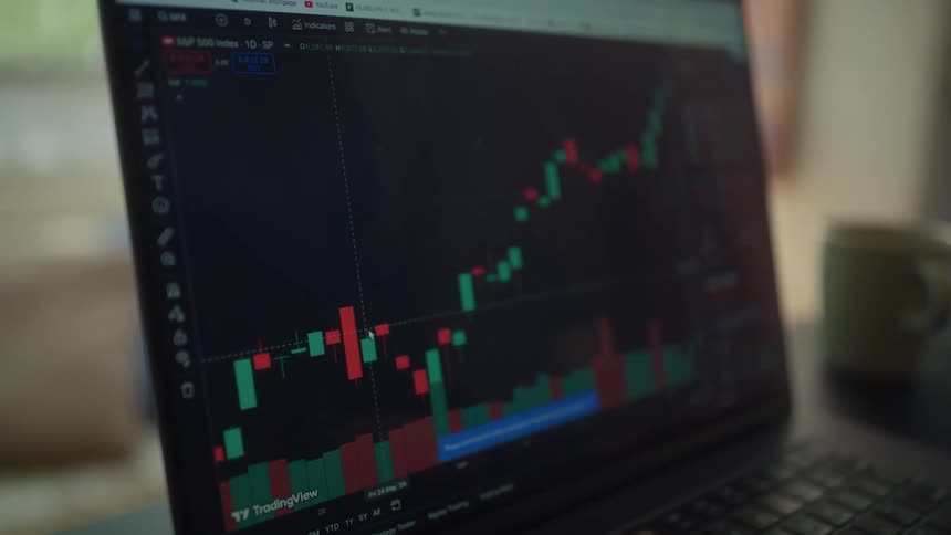 Person sits at a table at home and uses a computer displaying a graph related to trading