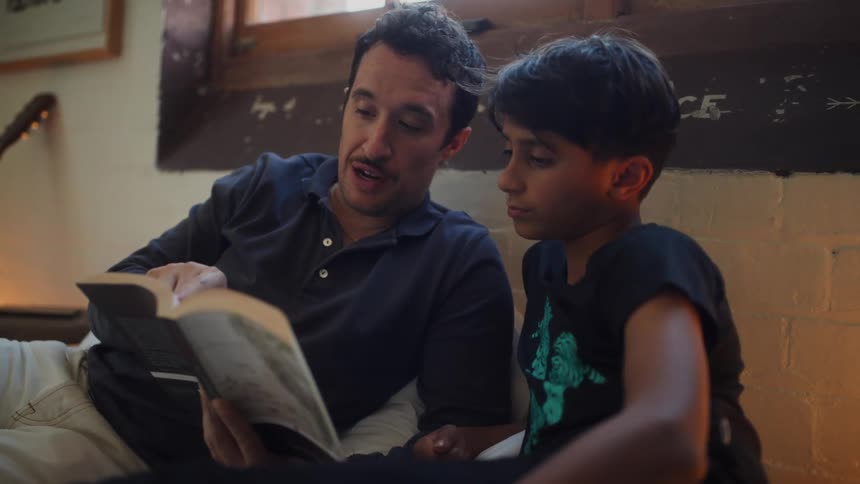 A father and son are seated closely together looking at a book the father is holding as he tells a story