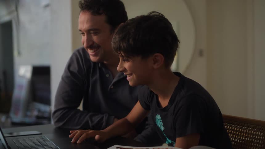 A father and son sit at a table at home with a laptop and homework sharing a smiling high five