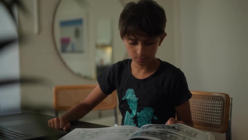 A boy sits at a table at home reading a book while browsing on a laptop