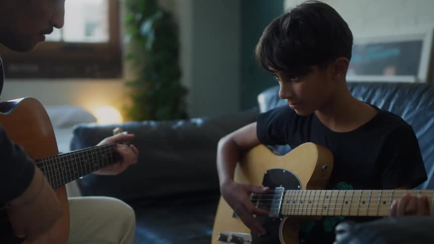 A father and son are playing guitars together in a living room with the son focused on playing an electric guitar while the father plays an acoustic guitar