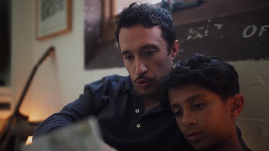 A father with a mustache and a dark blue shirt and son wearing a black t shirt are looking at a book together with the father appearing to read to the son who is listening attentively