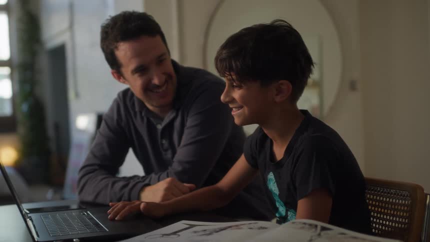 A father and son are looking together at a laptop screen with the father gesturing and the son focused on the display