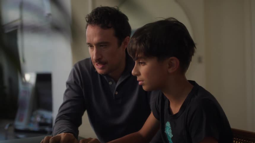 A father with a mustache and a dark shirt and son wearing a black t shirt are looking at laptop together both smiling