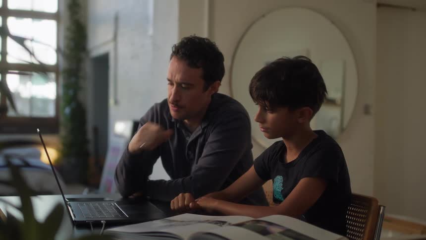 A father and son are seated at a table looking together at a laptop screen with an open book beside them