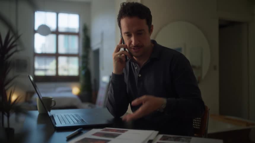 A man with curly dark hair sits at a desk talking on a cell phone while holding a document in a room with a laptop and a mug visible