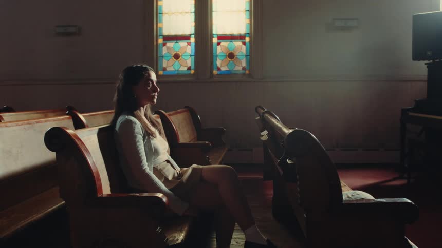 In a dimly lit church a young woman sits alone in a pew