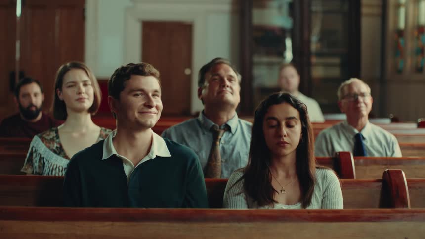 A man sitting next to a woman on a wooden pew inside a church scoots closer to her placing his hand over her shoulder and smiling while she appears indifferent