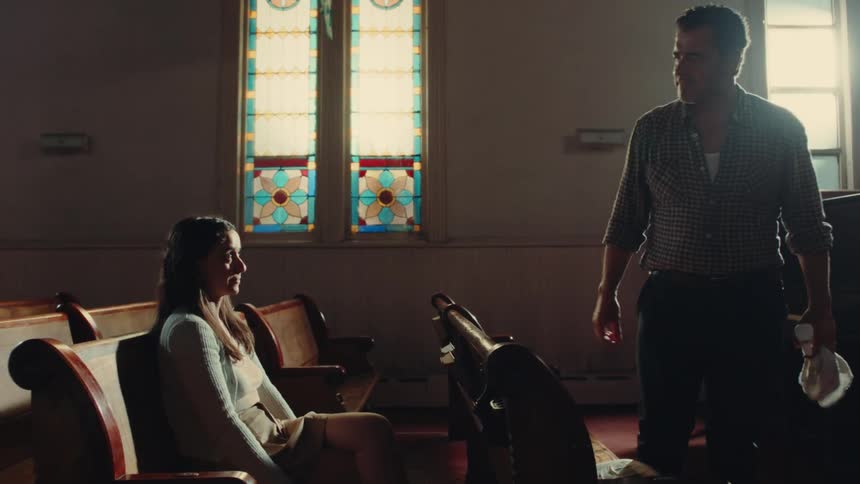 A younger woman sitting on a pew at church nods and shakes the hand of an older man standing there he pats her shoulder and points and she hangs her head