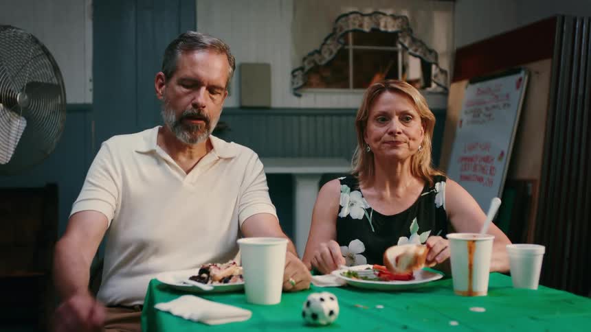 A man and a woman sit at a table with a green tablecloth displaying expressions of frustration and impatience while eating their meal