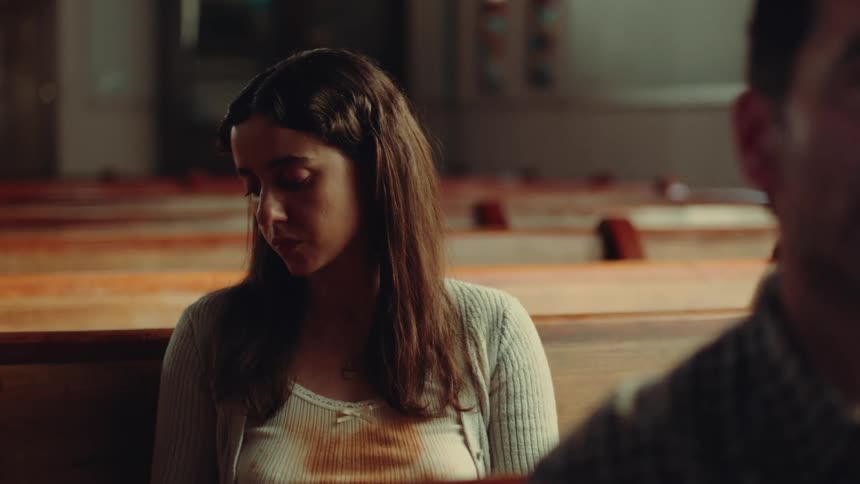 A woman in a stained shirt sits in a church pew behind another person appearing distracted