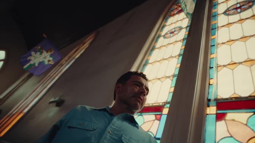 A man looks upward towards tall colorful stainedglass windows in a dimly lit church