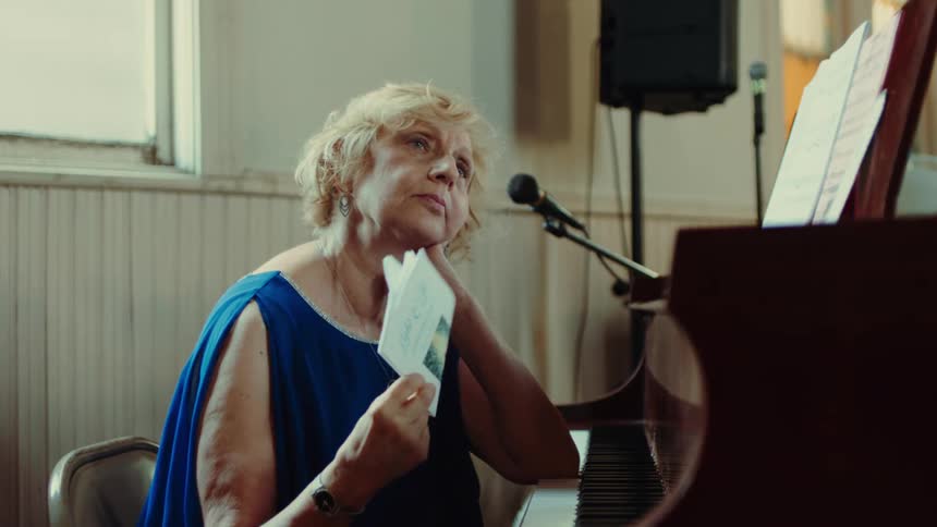 A woman sits at a piano in a church next to a microphone fanning herself with a pamphlet on a hot sunny day