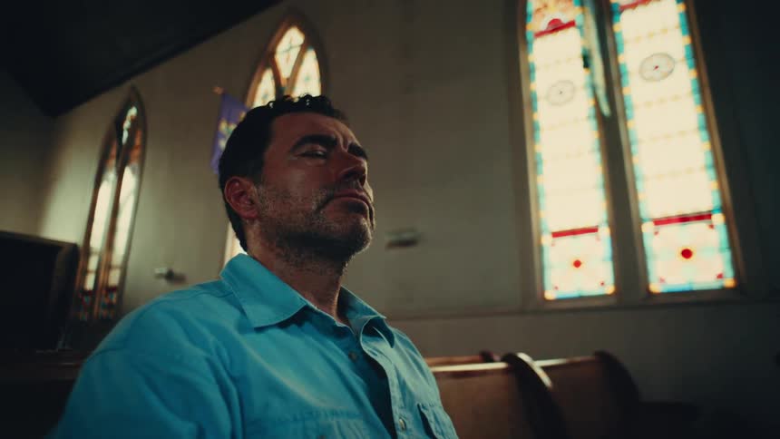 A man with his eyes closed sits in a church with stained glass windows visible in the background