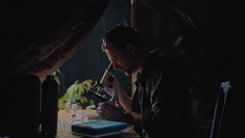 A man wearing glasses and gloves is intently using a microscope in a dimly lit setting with various vials and equipment scattered on the table around them