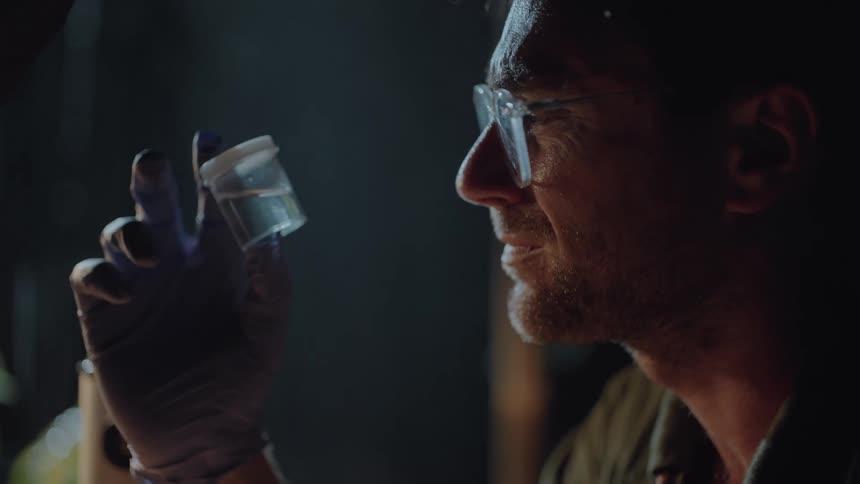 A man wearing glasses is carefully examining a small clear container held in his gloved hand