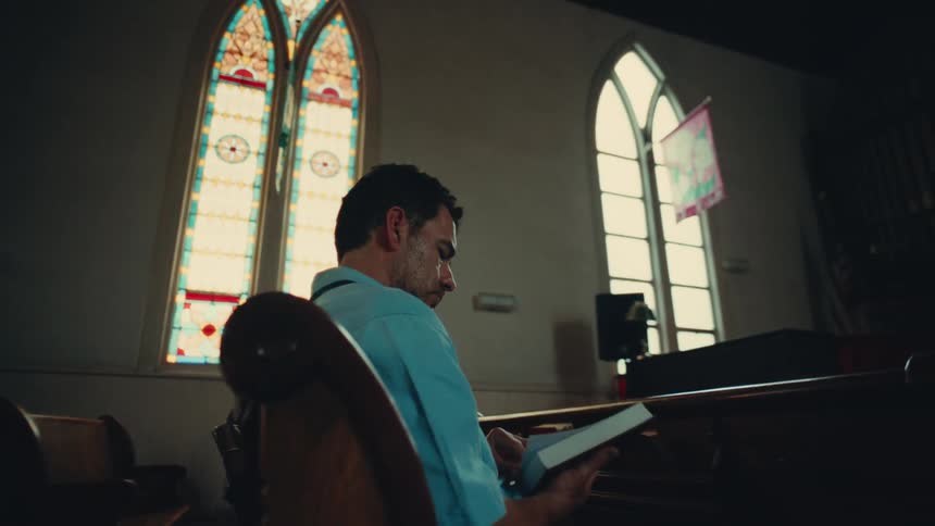 A man in a blue shirt is seated in a church pew reading a book with stained glass windows in the background