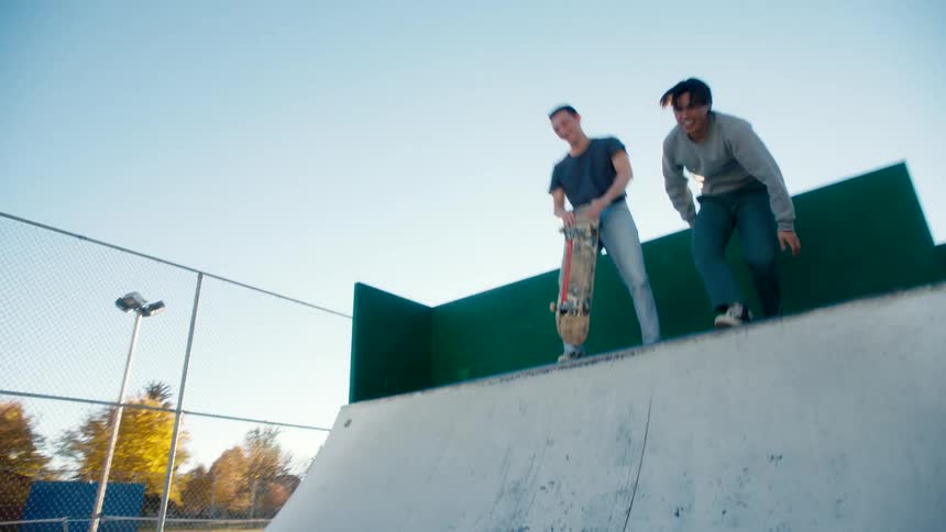 A teenager carrying a skateboard attempts to climb up a ramp at a skate park while their friends at the top playfully push him down in the morning