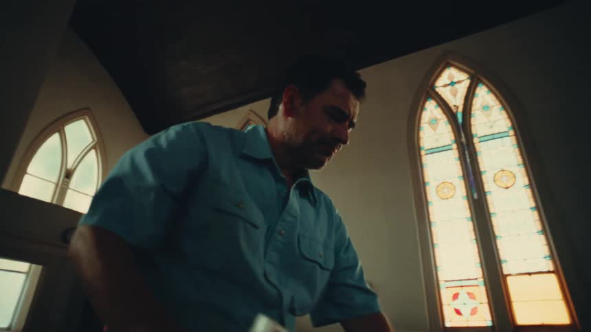 A janitor uses a rag to clean pews in a church with large glass windows
