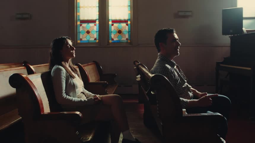 A man speaks while seated on a wooden pew in a church with stained glass windows as a woman behind him looks impatient