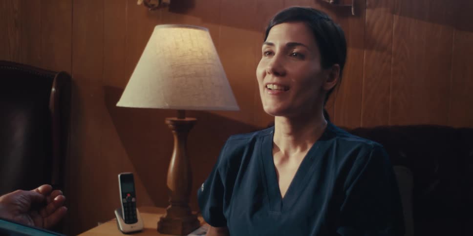 A healthcare worker wearing dark scrubs and nitrile gloves talks to an elderly patient