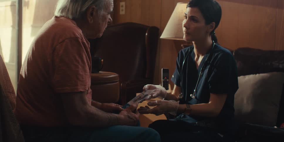 A nurse with a stethoscope tends to an elderly man sitting on a couch at home