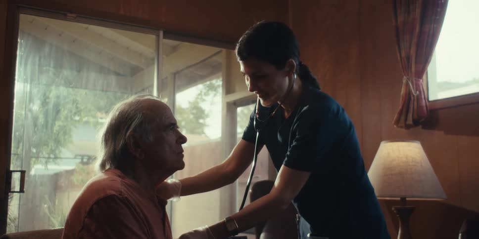 Nurse wearing blue scrub stands over an elderly man to listen to his heart beat the room has natural light and lamp light lighting up a light wooded paneled room
