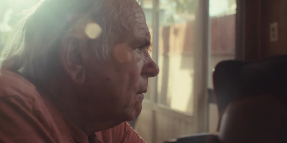 A female nurse with black hair wearing blue scrubs sits face to face with an elderly man she appears to be explaining something to him with a look of concern on her face as the elderly man listens intently