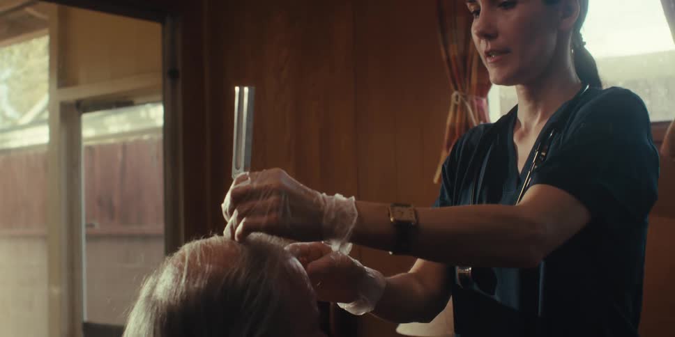 A healthcare professional wearing a dark scrub top gloves and a watch is examining the head of an elderly person with white hair