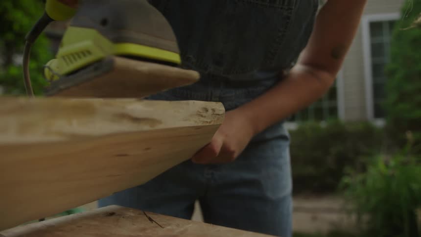 Female carpenter uses a power sander on a large plank of wood while standing in a residential yard on a summer day