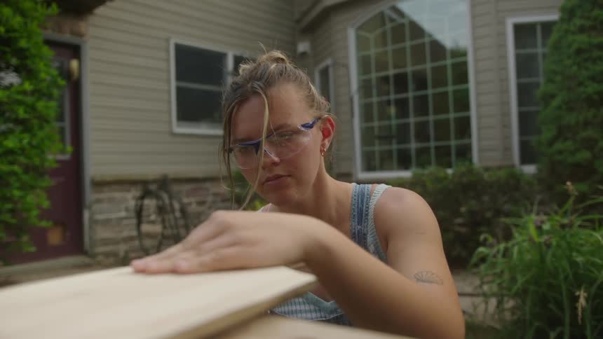 A woman kneels next to a wooden chopping board sanding it with a power tool in her backyard at home