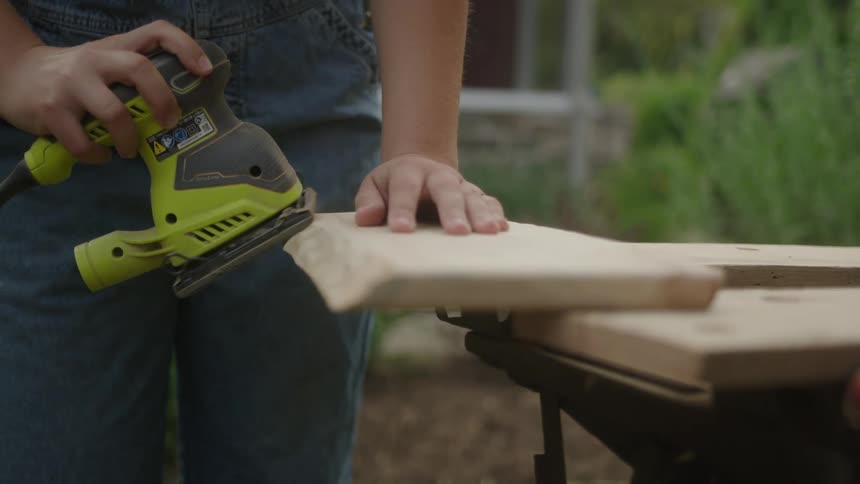 A closeup shows a persons hands sanding the edge of a plank outdoors