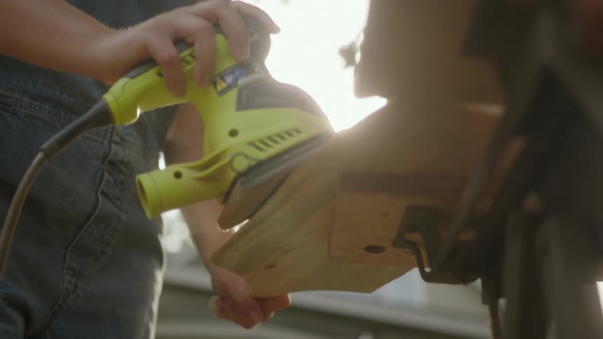 A person wearing a blue overall is sanding a piece of wood with a power tool at morning