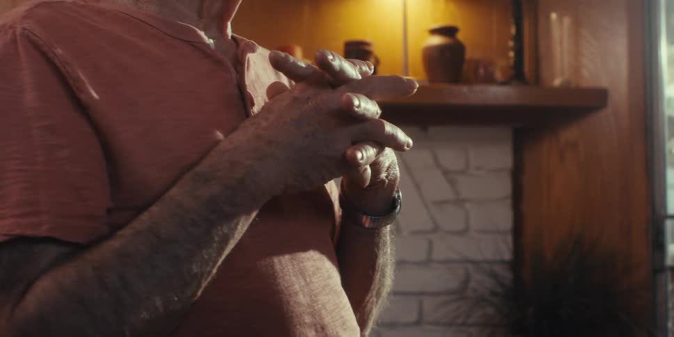 An elderly man clasps his hands to his chest with fingers intertwined as another person speaks to him