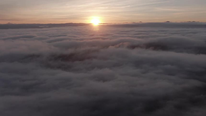 The sun sets above a vast expanse of clouds casting a soft warm glow across the misty landscape