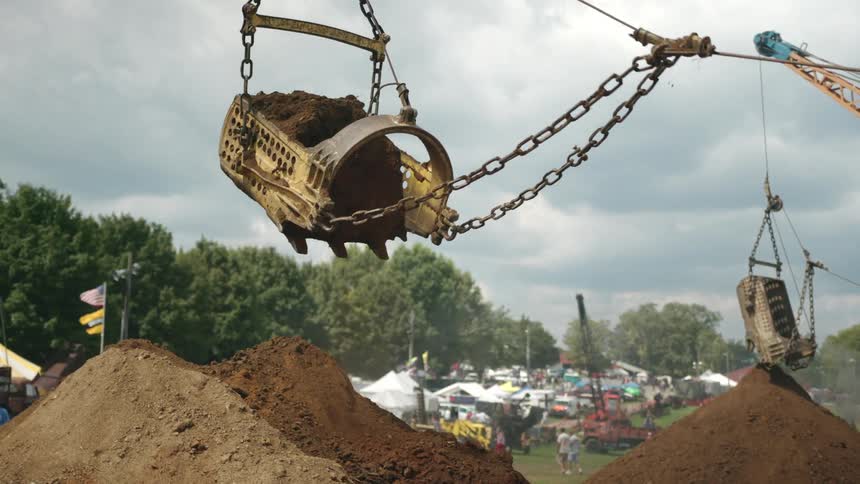 A large excavator lifts a bucket of dirt at a construction site with mounds of dirt in the foreground and a fairground with tents and people in the background