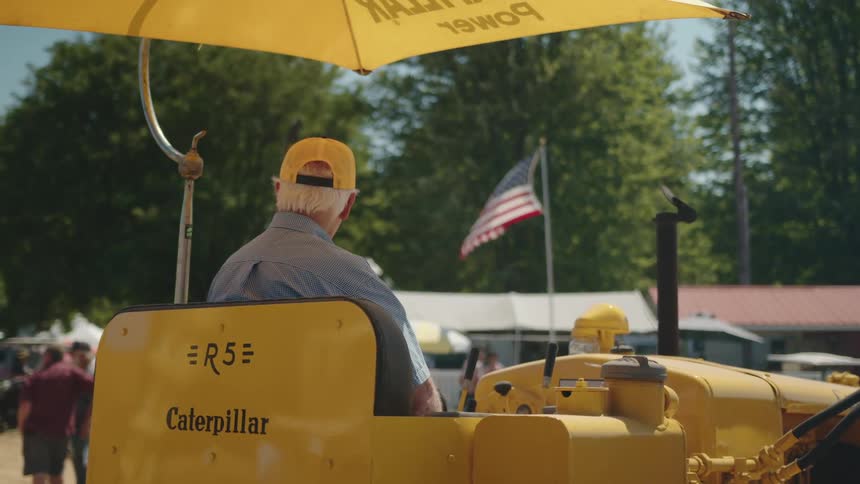 The back of an elderly man with white hair wearing a yellow hat sitting under a yellow umbrella on a yellow tractor at a fair with an american flag waving in the background